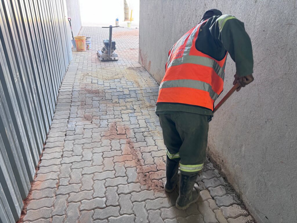 Worker sweeping sand over finished paving next to a plate compactor